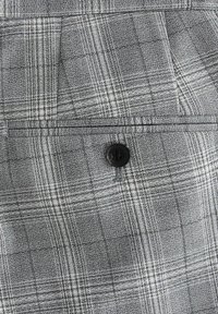 Tissu à carreaux gris présentant un tissage texturé avec de fines lignes et un détail en bouton noir sur une poche.
