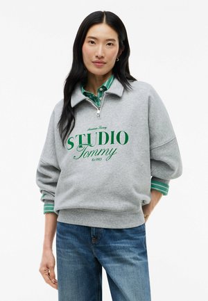 Femme aux longs cheveux noirs portant un sweat-shirt gris à fermeture éclair 1/4 avec l'inscription "Studio Tommy" par-dessus une chemise verte rayée et un jean bleu, debout, une main dans la poche.