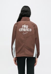 Καφέ αθλητικό μπουφάν με λευκές πλάγιες λεπτομέρειες, με το λογότυπο "nike athletics" και το σήμα Nike στον πίσω μέρος, κατασκευασμένο από ελαφρύ ύφασμα.