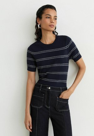 Kvinde med mørkt krøllet hår iført en navyblå kortærmet sweater med grå striber og mørke jeans med store forlommer, stående mod en hvid væg.