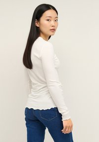 Top bianco a maniche lunghe in tessuto a coste con orlo arrotondato e polsini smerlati, abbinato a jeans blu. Texture liscia e design aderente.