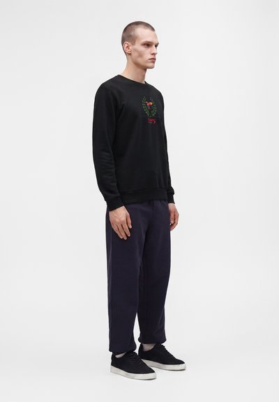 Jeune homme debout portant un sweat-shirt noir avec un logo, un pantalon ample foncé, des chaussettes blanches et des baskets noires sur un fond blanc uni.