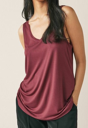 Kobieta w bezrękawniku bordowym z dekoltem w serek i czarnych spodniach, stojąca z rękami w kieszeniach na jednolitym tle.