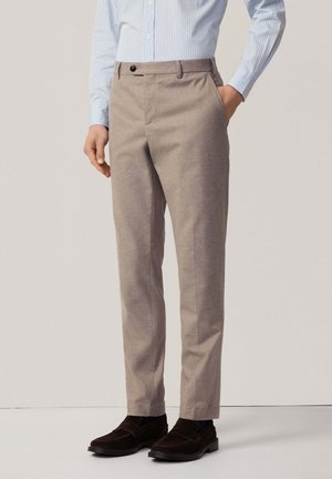 Mann trägt beige maßgeschneiderte Hose, ein hellblau gestreiftes Hemd und dunkelbraune Wildleder-Loafers mit einer Hand in der Tasche.