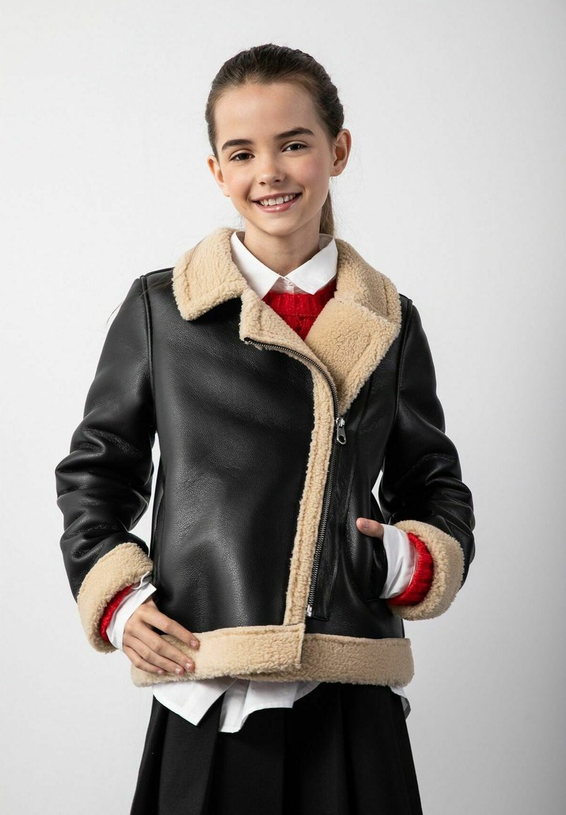 Czarna skórzana kurtka z beżowym kołnierzem i mankietami z owczej wełny, z zamkiem z przodu i dopasowanym krojem. Nosi się ją na białej koszuli i czerwonym swetrze.