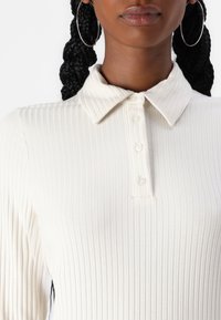 Frau mit einem weißen, gerippten Langarm-Polohemd mit vier Knöpfen und großen silbernen Creolen.