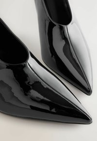 Zapatos de tacón de charol negro con punta afilada, acabado brillante y laterales abiertos, que cuentan con una textura suave y un sutil detalle de costura.