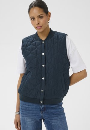 Vrouw die een marineblauwe doorgestikte bodywarmer met drukknopen draagt over een wit shirt met korte mouwen en een blauwe spijkerbroek, staand tegen een effen achtergrond.