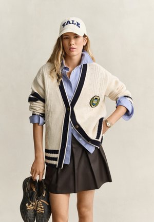 Jeune femme portant une casquette Yale et un cardigan crème, tenant une paire de baskets noires usées dans une main, vêtue d'une chemise bleue et d'une jupe plissée noire.