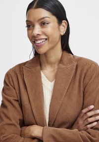 Brown corduroy blazer with a notched collar, worn over a cream knit sweater. The model has smooth, sleek hair and subtle earrings.