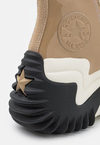 Beżowe sneakersy z błyszczącą cholewką, teksturowaną czarną gumową podeszwą i ciemną gwiazdką jako akcentem. Posiadają okrągłe logo oraz teksturowane detale.