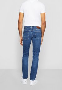 Hombre vistiendo jeans azules, camiseta blanca y zapatillas blancas de pie, de espaldas, contra un fondo gris liso.