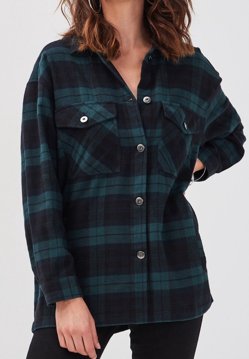 Femme portant une chemise à carreaux noire et vert foncé avec deux poches poitrine, debout avec une main sur la hanche, sur un fond uni.