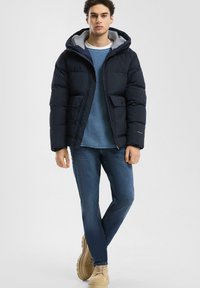 Mornarsko puffer jakna s sivo kapuco, z dvema sprednjima žepoma, v kombinaciji z svetlo modrim pletenim puloverjem in modrimi džinsom. Čevlji so svetlo rjavi.