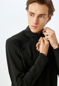 Top deportivo negro con cuello alto y cierre de cremallera, con mangas raglán y una textura suave. Diseño minimalista sin patrones.