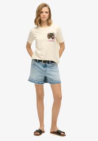 T-shirt en coton crème avec un motif de dragon et le texte "Superdry", associé à un short en denim bleu clair avec des bords effilochés. Sandales noires à enfiler.