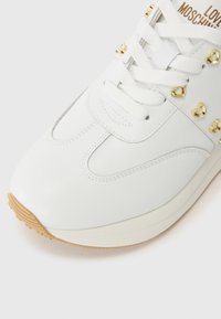 Sapatilhas brancas de couro com studs em forma de coração dourados, atacadores brancos planos e uma sola de borracha texturizada. Apresenta costuras contrastantes e uma biqueira arredondada.