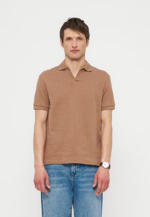 Mand iført en brun kortærmet ribstrikket polo shirt og blå jeans, stående mod en ensfarvet hvid baggrund.