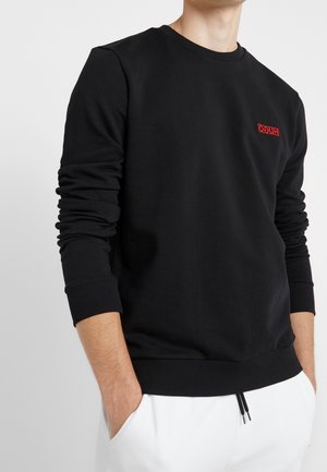 Mann trägt schwarzen Langarm-Sweatshirt mit rotem "HUGO"-Logo auf der Brust, Hände in den Taschen der weißen Hose, vor einfarbigem Hintergrund.