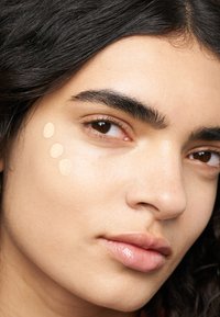 Un fond de teint liquide dans une teinte beige clair est appliqué en trois points sur une joue, mettant en valeur une texture de peau lisse et un éclat subtil.