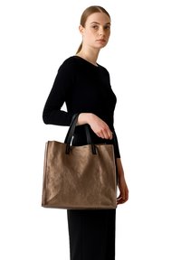 Borsa tote marrone strutturata con manici neri, caratterizzata da una forma rettangolare ampia e hardware minimal, tenuta da una persona con un top nero.