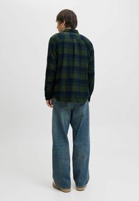 Person in einem grün-blauen karrierten Hemd, mit weitgeschnittenen blauen Jeans und beigen Schuhen, steht mit dem Rücken zu einer schlichten weißen Wand.
