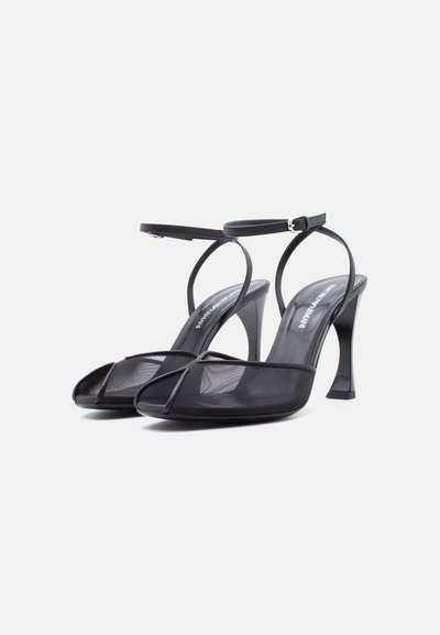 Sandales à talons hauts noires avec brides en mesh sur les orteils, fines brides à la cheville avec boucles, et talons évasés sur fond blanc.