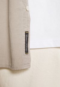 Camisa beige acanalada con cuello blanco, cierre de botones y una etiqueta negra con detalles dorados que dice "Marc O'Polo."