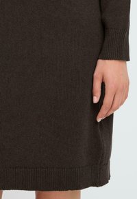 Brun strikket kjole med ribbestrikket kant og mansjetter. N�ærbilde viser tekstur og detaljer av stoffet. Hånd plassert nær sømmen på kjolen.