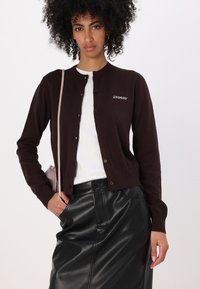 Mørkebrun cardigan laget av strikket stoff, med rund hals, knappelukking og brodert logo. Sammen med et svart skinnskjørt.