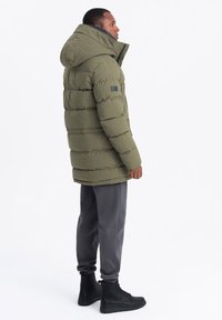 Chaqueta acolchada gruesa en color verde oliva con capucha, patrón de cuadrícula texturizado, cierre de cremallera y bolsillos laterales. Combinada con pantalones grises y botas negras.