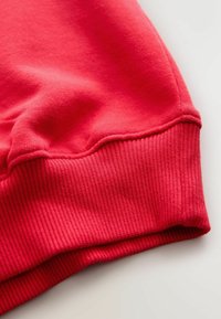 Manșetă roșie din bumbac a hanoracului, cu textură striată, având o suprafață exterioară netedă și detaliu cu cusătură la tranziția către manșetă.