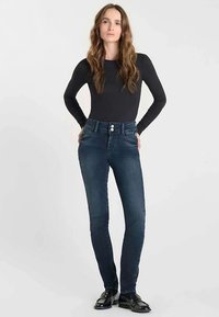 Jean skinny bleu foncé en denim avec une taille haute, deux boutons à l'avant, cinq poches et une texture lisse. Porté avec un haut à manches longues noir.
