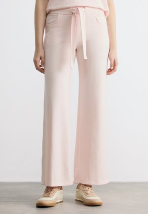 Osoba oblečená v svetloružových širokých nohaviciach s opaskom previazaným v páse a béžovými teniskami, stojaca na sivom podlaží.