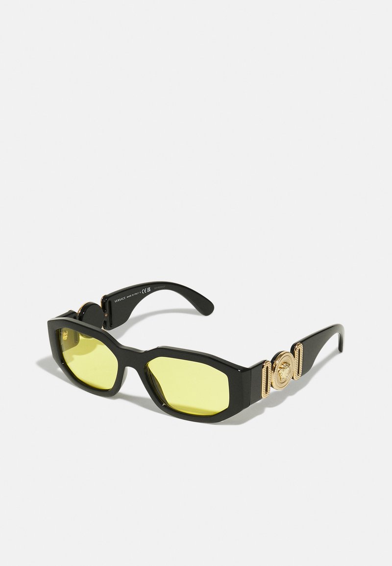 Óculos de sol pretos com lentes amarelas, design angular, acentos de hardware dourado proeminentes nas hastes e estruturas de plástico elegantes.