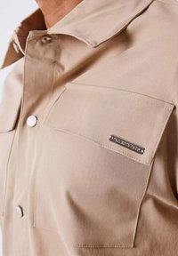 Camicia beige con bottoni, caratterizzata da un colletto a punta, due tasche sul petto e un'etichetta metallica, realizzata in tessuto liscio con una leggera lucentezza.
