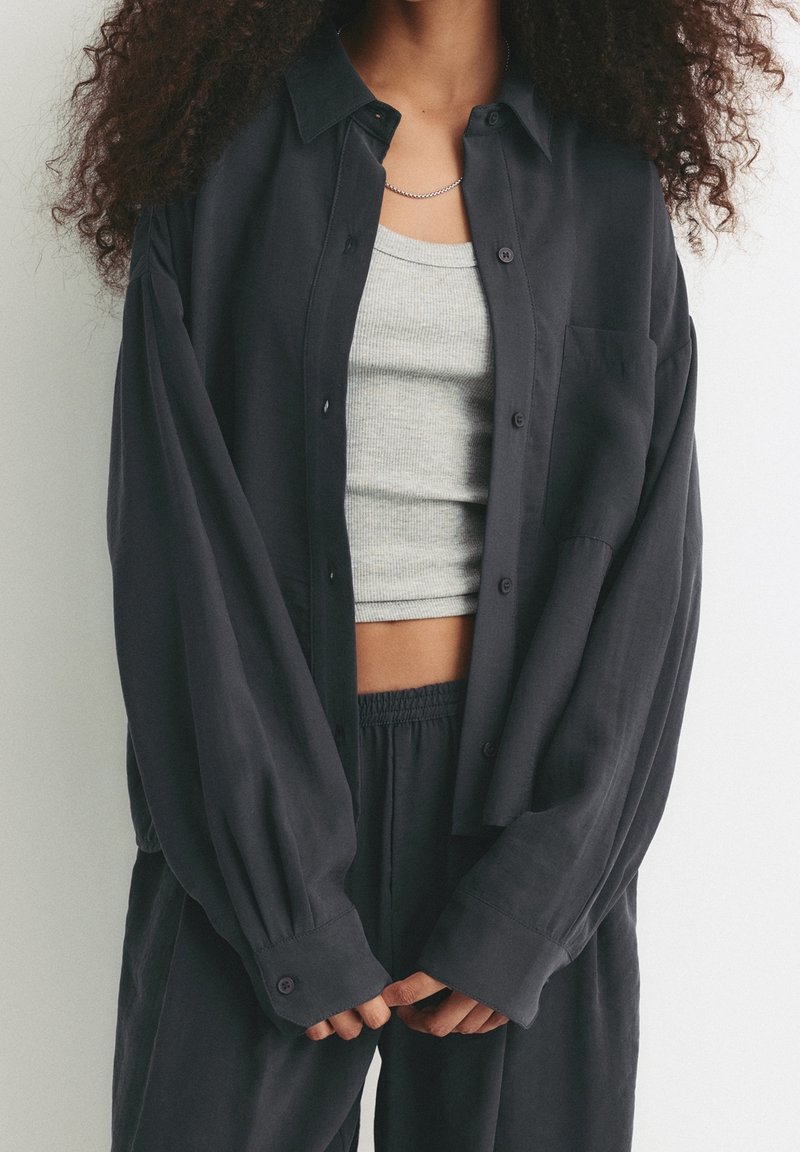 Personne portant une chemise noire ample boutonée par-dessus un crop top gris et un pantalon noir assorti, debout devant un fond clair uni.