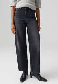Pantaloni larghi in denim nero con vita alta, leggero lucido e tasche frontali. Abbinati a stivaletti aderenti. La texture appare liscia.