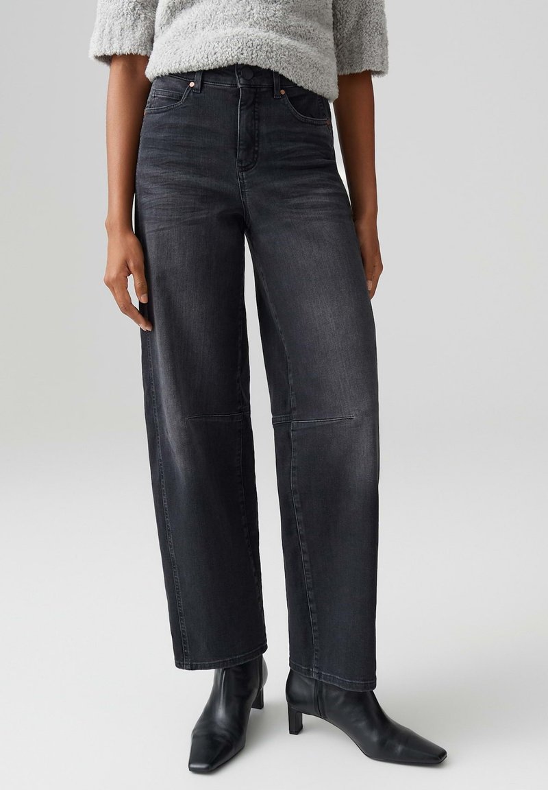 Pantaloni larghi in denim nero con vita alta, leggero lucido e tasche frontali. Abbinati a stivaletti aderenti. La texture appare liscia.