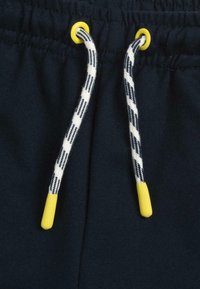 Pantalons de survêtement bleu marine avec un cordon de serrage aux embouts jaunes et un motif tressé en blanc et bleu marine. Tissu doux et texturé avec une taille élastique.