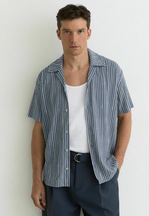 Mand iført blå og hvide lodrette stribede kortærmede skjorte over en hvid tanktop, med mørke bukser og hånden i lommen, mod en hvid baggrund.