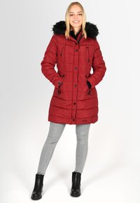Red puffer coat with fur-lined hood, front snap buttons, patch pockets, and visible stitching details. Worn over gray jeans and black boots.