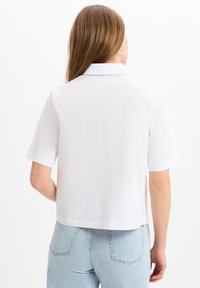 Witte short-sleeve shirt met kraag, gladde textuur, cropped ontwerp en rechte zoom. Draagt met lichtblauwe high-waisted jeans.