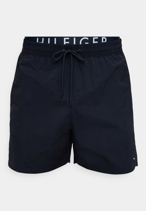 Marineblå herrebadeshorts med elastisk midje, sentral snor, logobånd på innsiden og liten flaggemblem nær kanten.