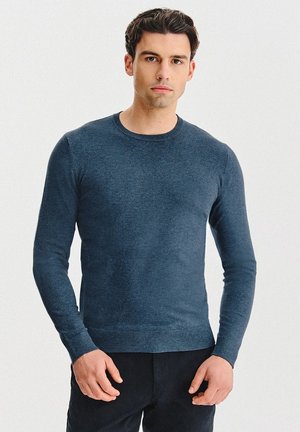 Granatowy sweter z długim rękawem, wykonany z miękkiej, teksturowanej dzianiny. Posiada okrągły dekolt i dopasowane mankiety, zestawiony z ciemnymi spodniami.