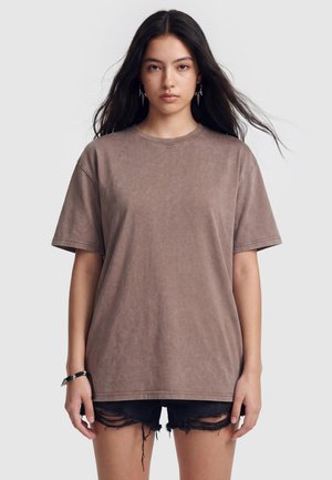 Ung kvinde med langt mørkt hår iført en løs brun t-shirt og sorte revnede shorts, står med et neutralt udtryk.