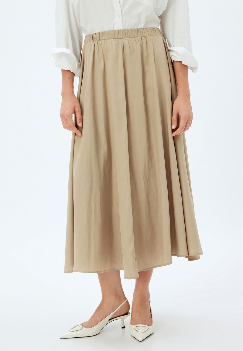 Jupe maxi beige avec une taille élastique, en tissu léger et au design fluide. Portée avec des chaussures à talons ouverts blanches ornées d'un accent décoratif.