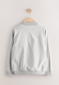 Szara bluza z teksturowanej tkaniny, z prążkowanymi mankietami i kołnierzem oraz prostym krojem. Charakteryzuje się prostym i czystym designem. Zawieszona na drewnianym wieszaku.