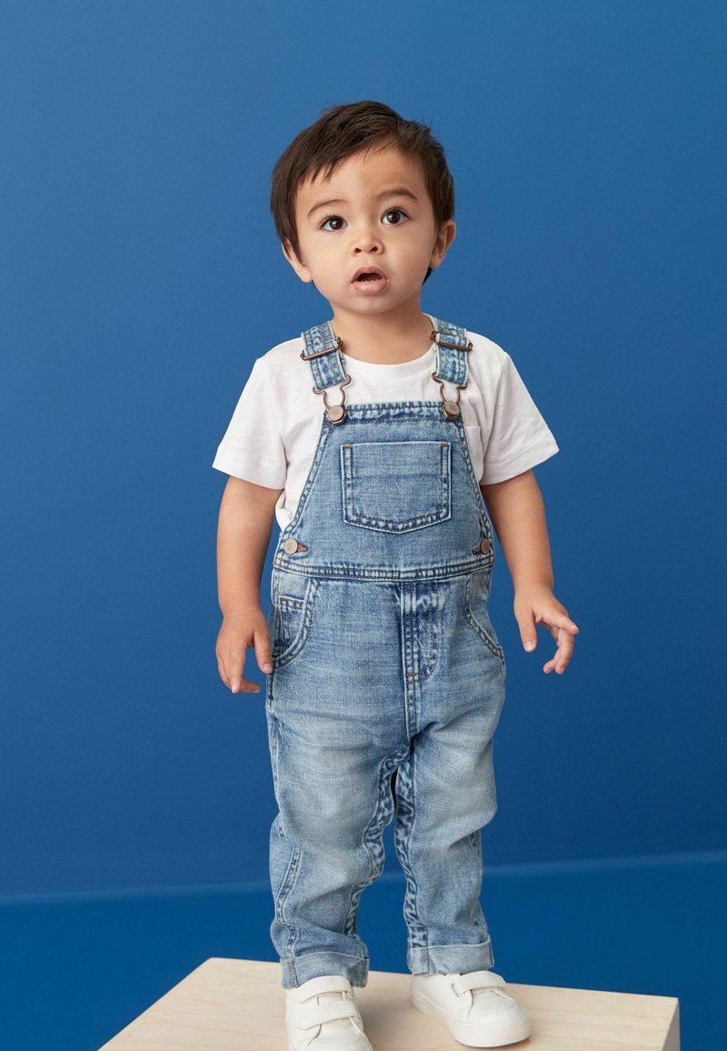 Denim overalls med justerbare stropper, lys blå vask, frontlomme og opkrøllede kanter; parret med en hvid t-shirt og hvide sneakers.