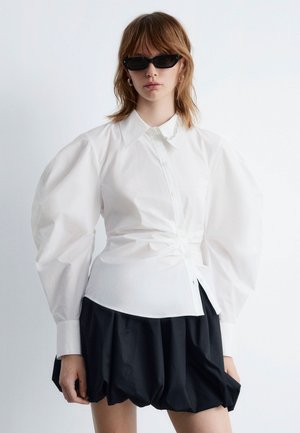 Weißes Hemd mit voluminösen Ärmeln, kombiniert mit einem schwarzen, puffigen Rock. Model trägt schwarze Katzenaugen-Sonnenbrillen und eine Silberkette.